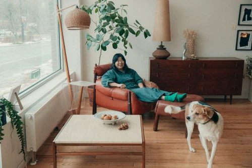 Woman in blue dress lounges in red chair with dog in bright, modern living room, featuring large window, lamp and wooden floors.