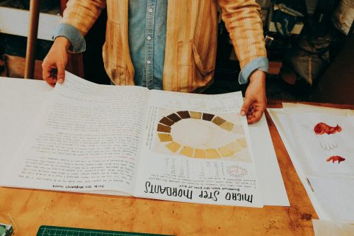 Close-up of an open book with handwritten text and a color chart, held by a person wearing a blue shirt and striped jacket.