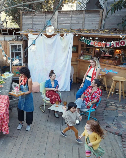 Outdoor gathering with a group of women and children on a wooden deck near a bar, featuring casual attire, string lights, and a festive atmosphere.