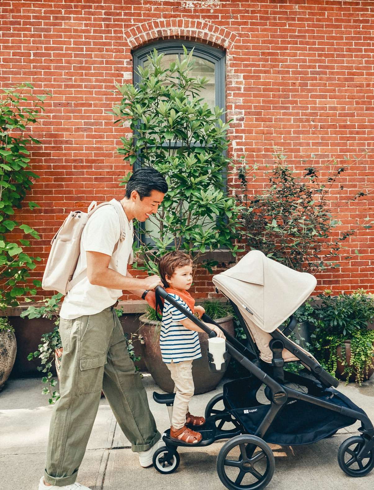 Father walks with his child on a stroller attachment on a city street, plants and brick building in background. 