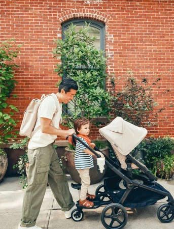 Father walks with his child on a stroller attachment on a city street, plants and brick building in background. 
