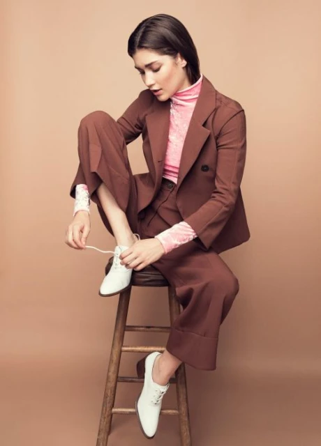 Brunette woman in brown suit seated on a wooden stool, tying the laces of her white shoes against a tan studio backdrop.