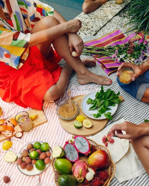 People at a poolside picnic with vibrant summer fruits, cocktails, and colorful textiles create a relaxed outdoor scene.