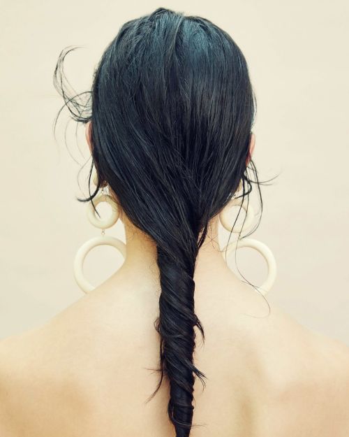 Rear view of a woman with long dark hair twisted into a vertical braid, wearing large cream hoop earrings against a neutral background.