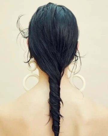Rear view of a woman with long dark hair twisted into a vertical braid, wearing large cream hoop earrings against a neutral background.