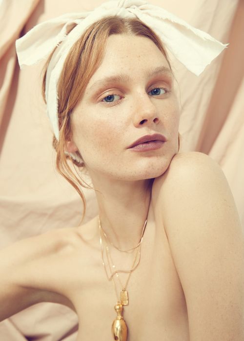 Close-up portrait of a woman with red hair wearing a white headband and layered gold necklaces against a neutral fabric background.