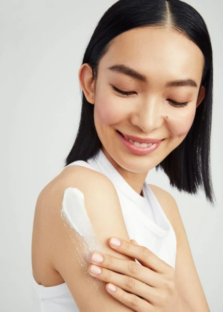 Smiling Asian woman applies white cream to her upper arm in a beauty and skincare product shot on a plain white background.