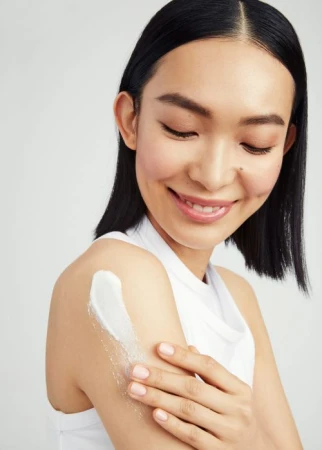 Smiling Asian woman applies white cream to her upper arm in a beauty and skincare product shot on a plain white background.