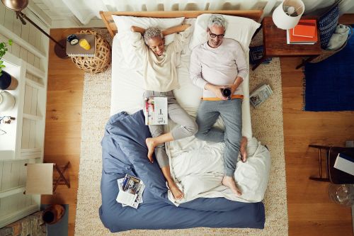 Overhead shot of a casually dressed mature couple relaxing together in bed, bathed in natural light from a nearby window.