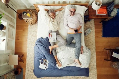 Overhead shot of a casually dressed mature couple relaxing together in bed, bathed in natural light from a nearby window.