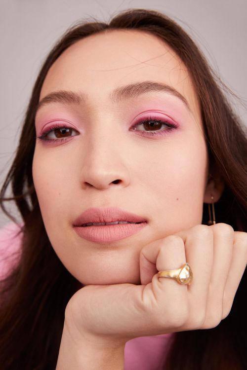 Close-up of a woman with pink eye shadow, light pink lipstick, and dark brown hair resting her chin on her hand while wearing a ring.