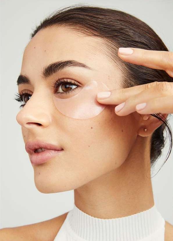Close-up of an adult woman applying a light pink under-eye mask, showcasing skincare routine and cosmetic application against a neutral backdrop.