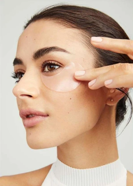 Close-up of an adult woman applying a light pink under-eye mask, showcasing skincare routine and cosmetic application against a neutral backdrop.