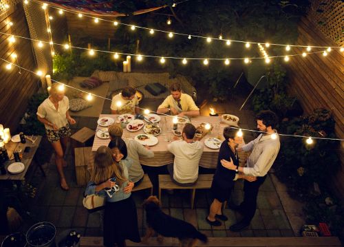 Overhead view of group of people dining outdoors in the evening, illuminated by string lights and candles on wooden patio.