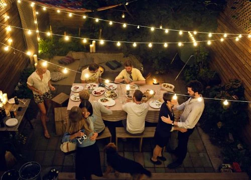 Overhead view of group of people dining outdoors in the evening, illuminated by string lights and candles on wooden patio.