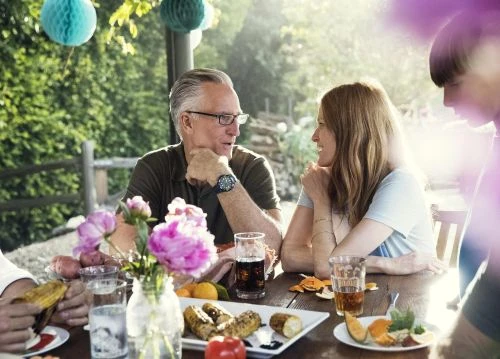 Family gathers outdoors at a table covered with food and drinks for a meal together, enjoying a sunny day in a garden setting.