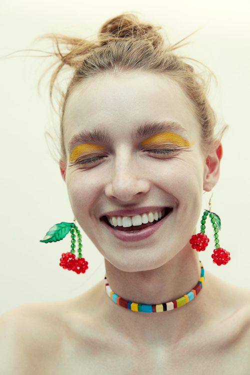 Smiling young adult female with fair skin and a messy bun, wearing cherry earrings, yellow eyeshadow, and a colorful beaded necklace.