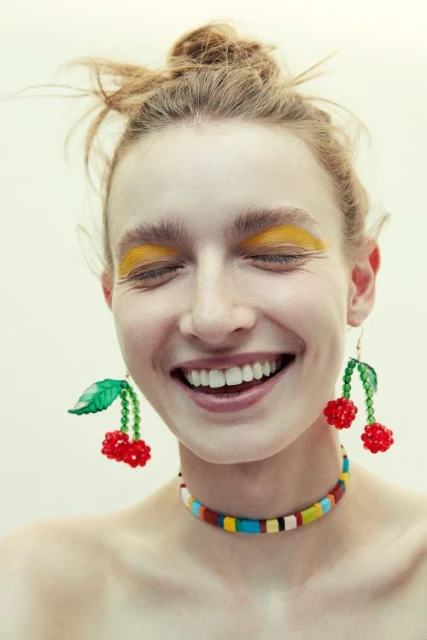 Smiling young adult female with fair skin and a messy bun, wearing cherry earrings, yellow eyeshadow, and a colorful beaded necklace.