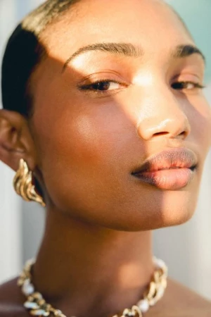 Close-up portrait of a woman wearing gold earrings and a chunky gold and pearl necklace under warm sunlight