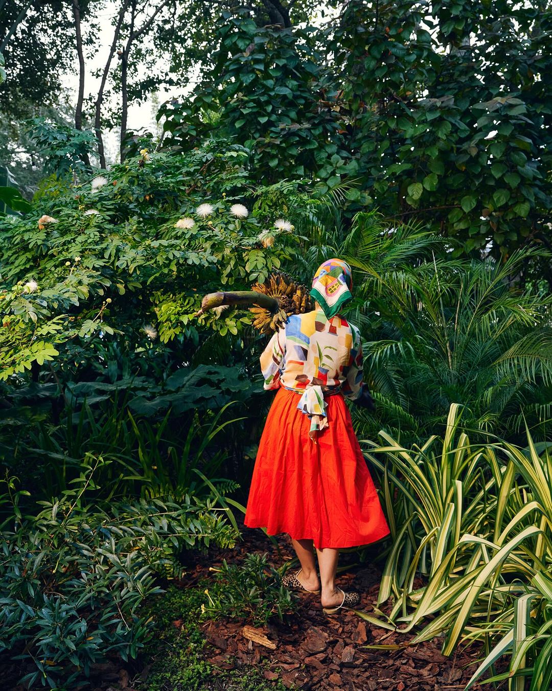 Woman in a colorful blouse and red skirt stands in lush greenery with various plants and trees in a natural outdoor environment.