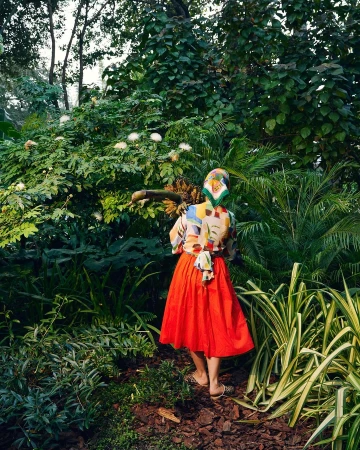 Woman in a colorful blouse and red skirt stands in lush greenery with various plants and trees in a natural outdoor environment.