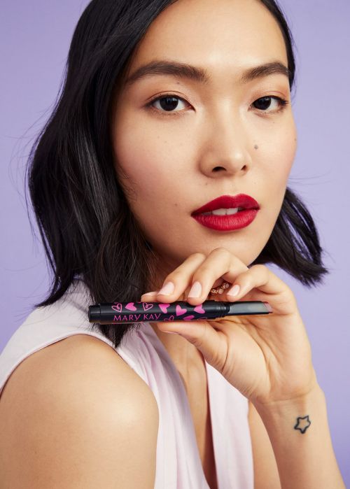 Asian female holding makeup product with pink heart design, red lipstick, light purple background, head and shoulders portrait.