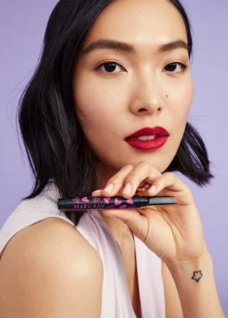 Asian female holding makeup product with pink heart design, red lipstick, light purple background, head and shoulders portrait.