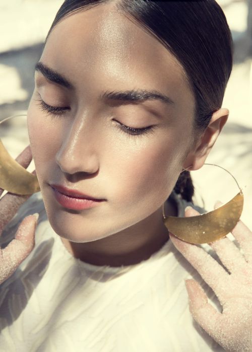 Portrait of an adult woman with closed eyes wearing large gold hoop earrings and a light top in soft light, showing a beauty and fashion aesthetic.