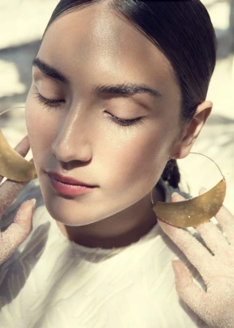Portrait of an adult woman with closed eyes wearing large gold hoop earrings and a light top in soft light, showing a beauty and fashion aesthetic.