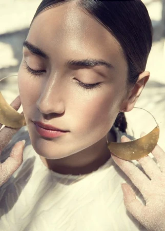 Portrait of an adult woman with closed eyes wearing large gold hoop earrings and a light top in soft light, showing a beauty and fashion aesthetic.
