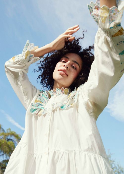 Woman in white dress with ruffled collar and cuffs raises arms against blue sky in low-angle shot, trees visible in the background.