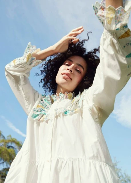 Woman in white dress with ruffled collar and cuffs raises arms against blue sky in low-angle shot, trees visible in the background.