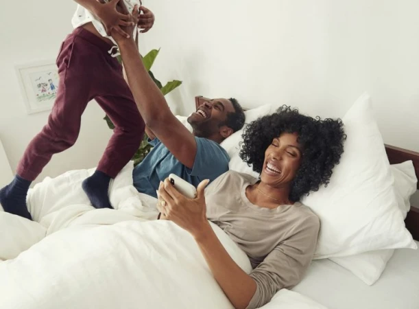 Black family laughing in bed with a woman holding a smartphone and a child playing above