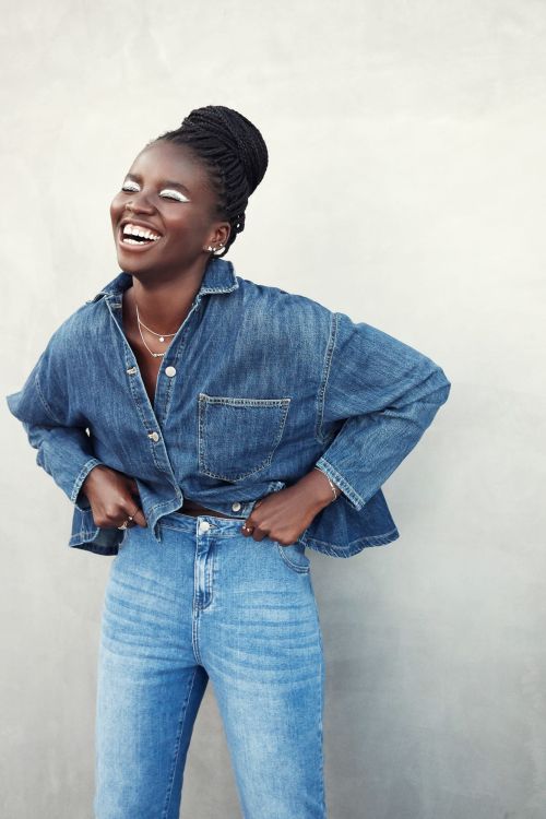 Smiling Black woman wearing a denim button-down shirt and high-waisted jeans against a plain grey concrete wall.