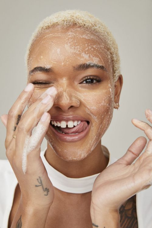 Close-up of a smiling woman with blonde hair and soap on her face, winking and sticking her tongue out in a beauty shot.