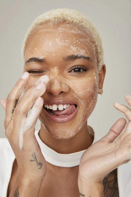 Close-up of a smiling woman with blonde hair and soap on her face, winking and sticking her tongue out in a beauty shot.