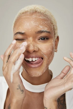 Close-up of a smiling woman with blonde hair and soap on her face, winking and sticking her tongue out in a beauty shot.
