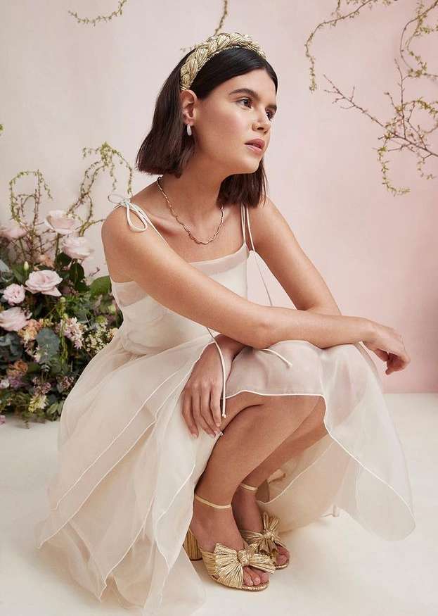 A woman with brown hair poses in a light dress and straw headpiece. She is in a studio with a light-pink backdrop and flowering branches.