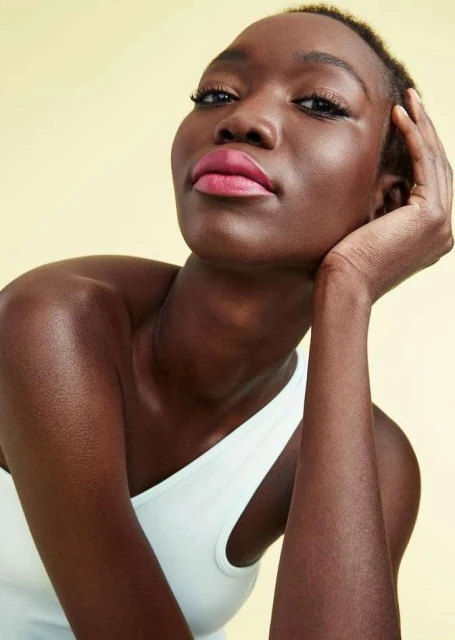 Portrait of a dark-skinned woman with her hand near her face, wearing a white top against a pale yellow background, looking at the camera.