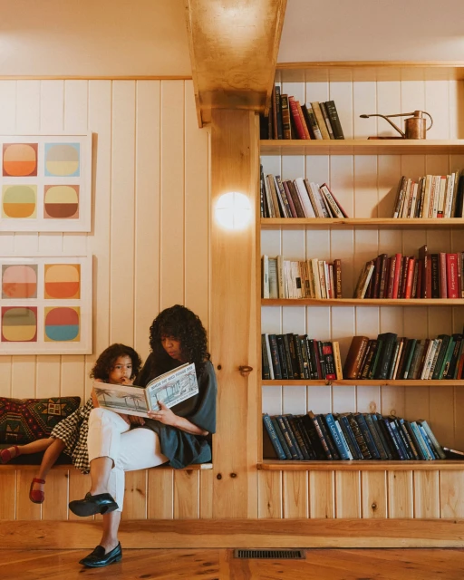 A woman and child sitting indoors reading a book near a bookshelf, with warm, inviting lighting and light wood panelling.
