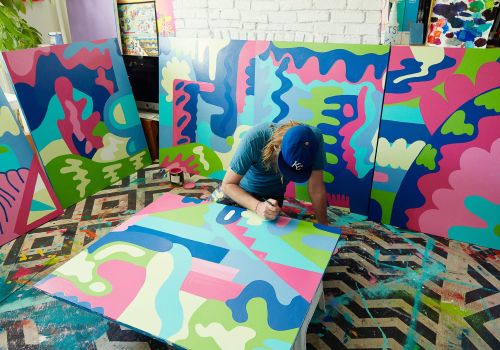 Artist in studio creating colorful abstract painting surrounded by finished artwork, wearing baseball cap, vibrant colors, patterned floor.