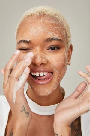 Close-up portrait of a smiling young woman with bleached-blonde hair, applying foamy cleanser to her face with her hands, winking.
