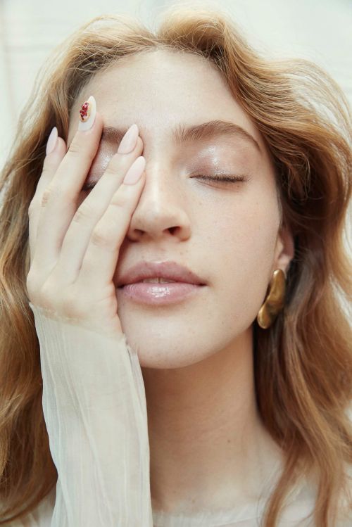 Close-up portrait of a young woman with light skin, wavy red hair, and neutral makeup, covering one eye with her manicured hand.