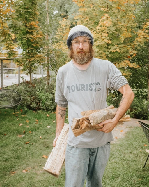 Adult male with a beard and knit hat outdoors, carrying firewood. Trees in the background and a casual outfit.