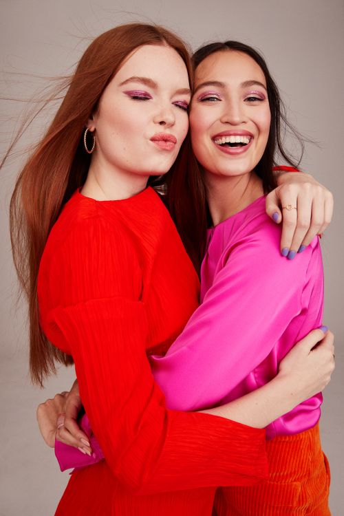 Two women embrace indoors wearing brightly colored tops; one in red with auburn hair poses puckered lips, the other smiles in hot pink.