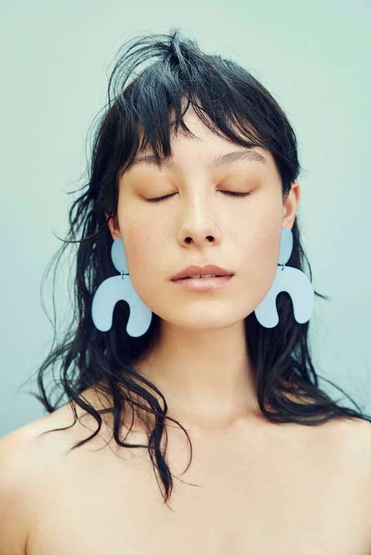 Woman with eyes closed wearing large light-blue earrings against a light-blue background in a studio portrait with soft lighting.