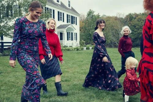 Several women and a toddler walk on the lawn outside a white house on a cloudy day, showcasing floral dresses and fall fashion.