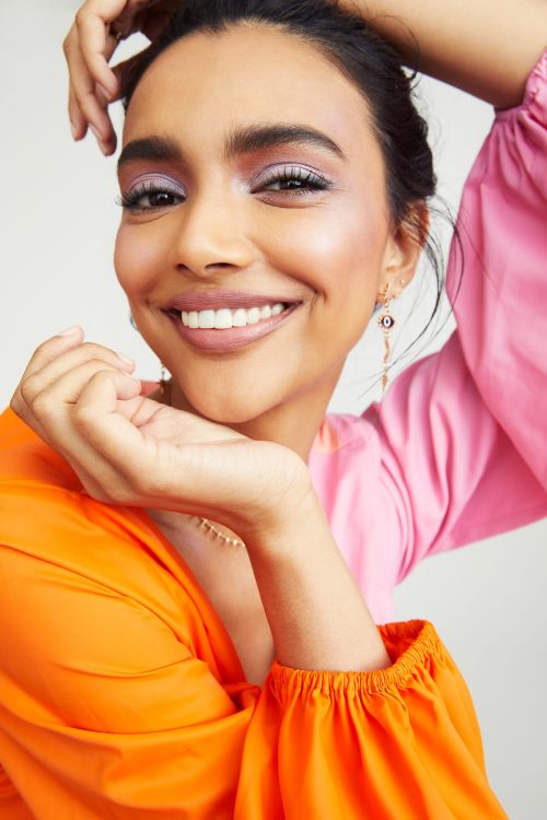 Close-up shot of a smiling woman with lavender eyeshadow, wearing a colorful top, and gold earrings against a white background.