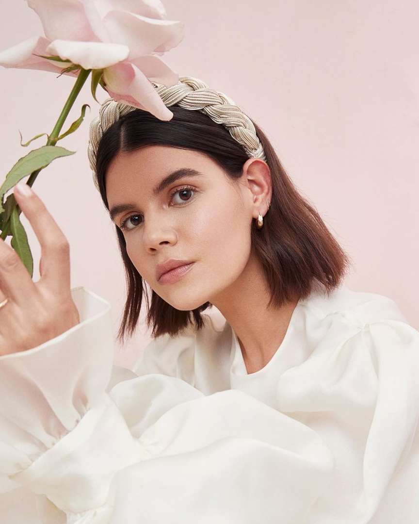 Close-up of an adult woman with a braided headband holding a pink flower against a light pink background in an indoor setting.