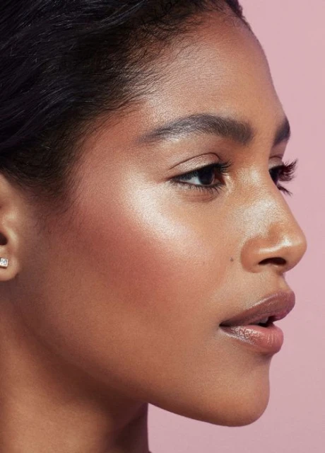 Close-up of a young woman with highlighted skin and glossy lips in front of a light pink background; beauty shot for the cosmetics industry.